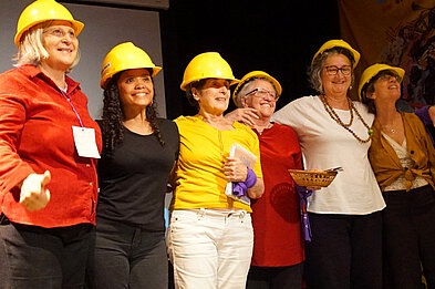 Hay cinco mujeres con cascos amarillos de construcción sobre el escenario, abrazadas y posando hacia adelante todas. Se ven felices.