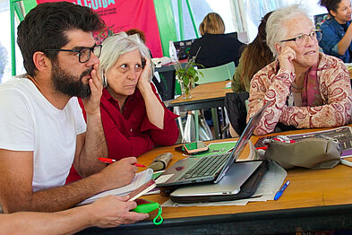 Hay tres personas sentadas en una mesa, de izquierda a derecha, un muchacho joven de lentes, a su lado dos señoras mayores. Escuchan con atención y miran en la misma dirección. Sobre la mesa hay una computadora y materiales para trabajar como papel y marcadores de colores.